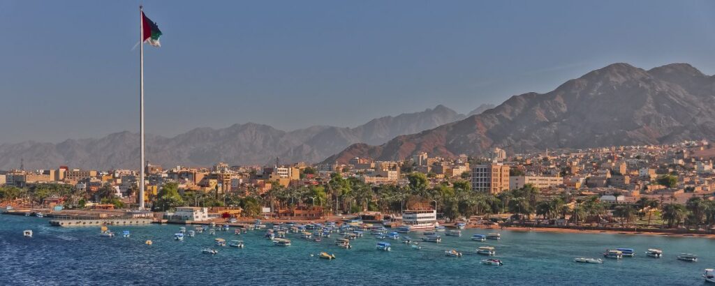 Coastal city waterfront with a tall flagpole, boats in a turquoise harbor, palm trees, and a distant mountain range under a clear sky.