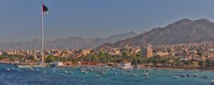 Coastal city waterfront with a tall flagpole, boats in a turquoise harbor, palm trees, and a distant mountain range under a clear sky.