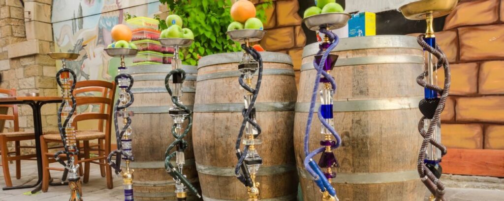 Row of tall hookahs with colorful hoses and fruit bowls on top, lined up in front of wooden barrels at an outdoor cafe area