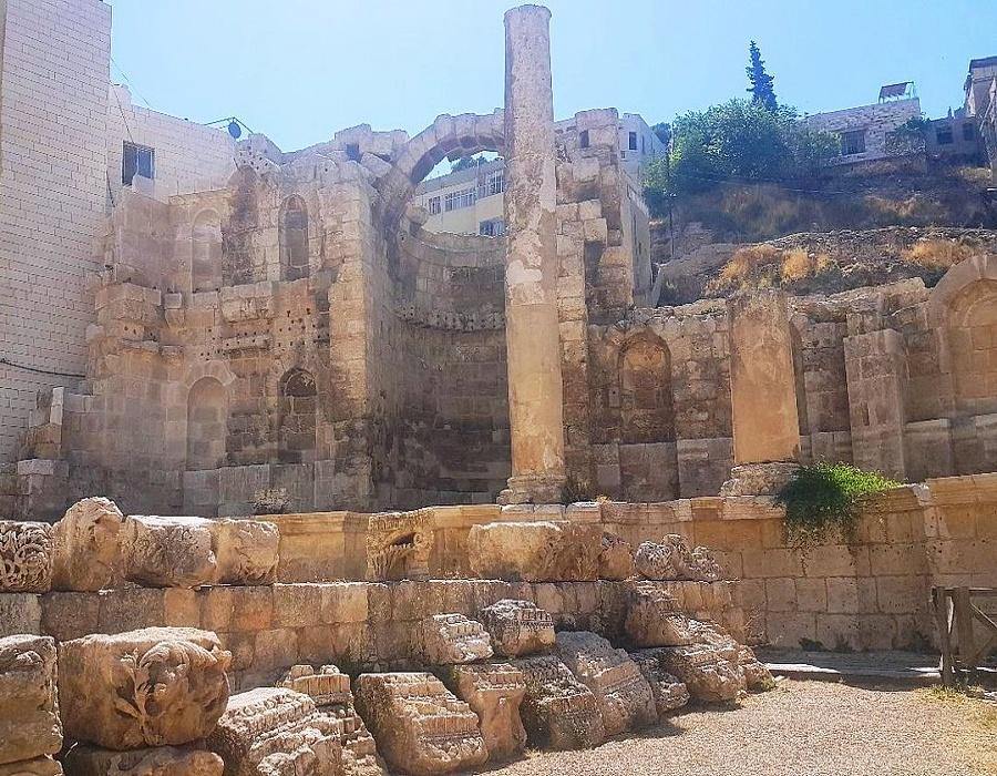 Ancient stone ruins with a tall central column and archways, scattered blocks on the ground under a bright sky.