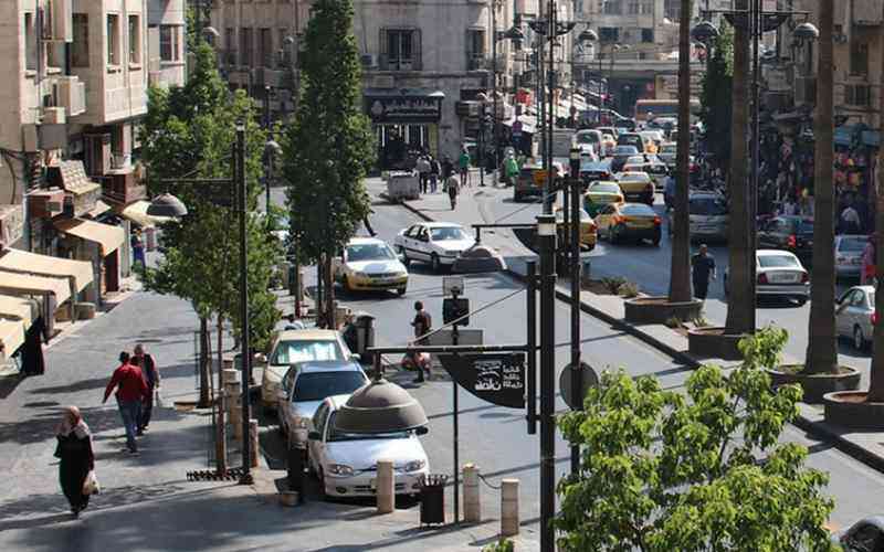 Busy urban street scene with cars and taxis, pedestrians walking along sidewalks, and trees lining the road. storefronts and people visible in the background.