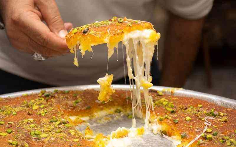 Hand lifting a slice of cheesy bread with gooey mozzarella strings from a large pan of spicy sauce in a metal tray.