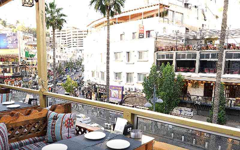 | Ordonna Guide Outdoor restaurant balcony with tables and cushions overlooking a palm-lined city street with shops and pedestrians.