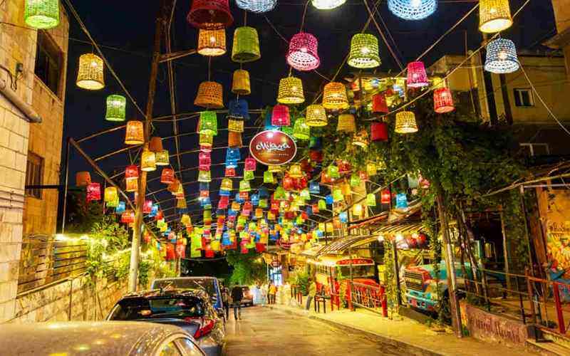 Night street scene with colorful hanging lanterns strung overhead, cars parked along the curb and pedestrians under festive lights at night.