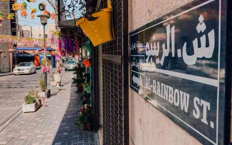 Colorful narrow street with hanging decorations, potted plants, and a storefront sign reading 'AL-RAINBOW ST.' on the right side of the image.