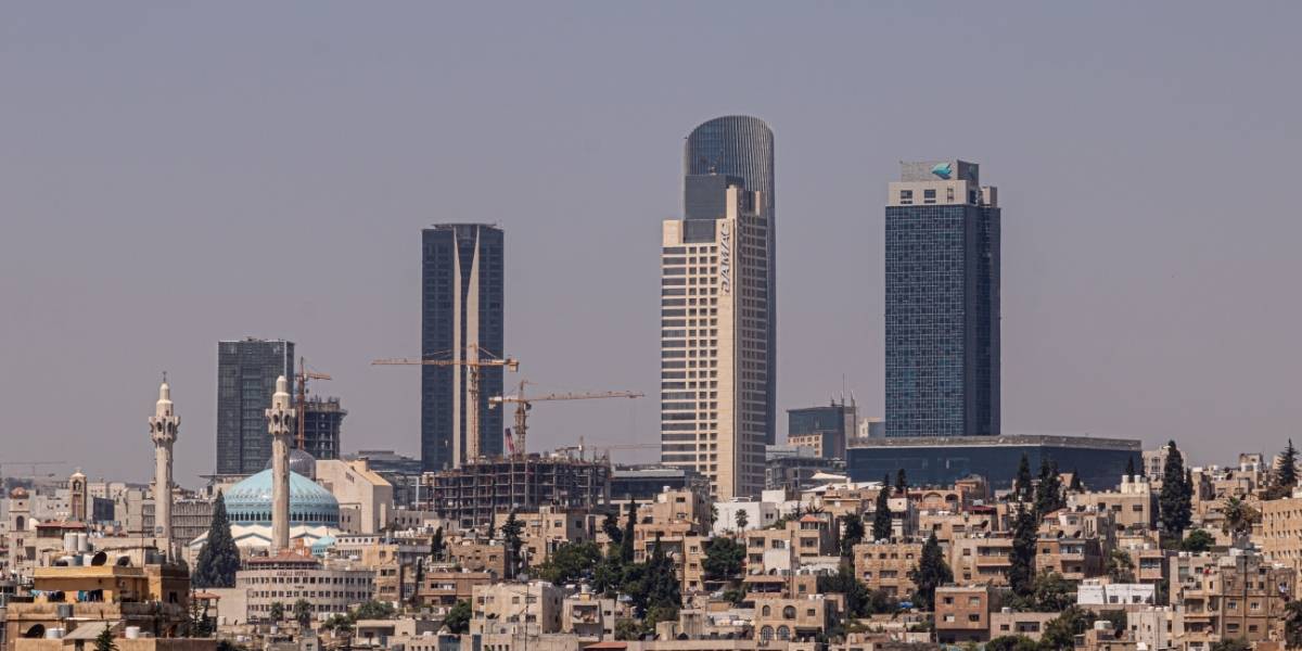 City skyline with tall modern towers and construction cranes rising above a dense low-rise district, under a clear sky.