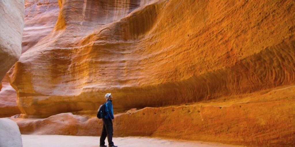 Hiker wearing a blue jacket and backpack stands in a narrow orange sandstone canyon of layered rock formations.
