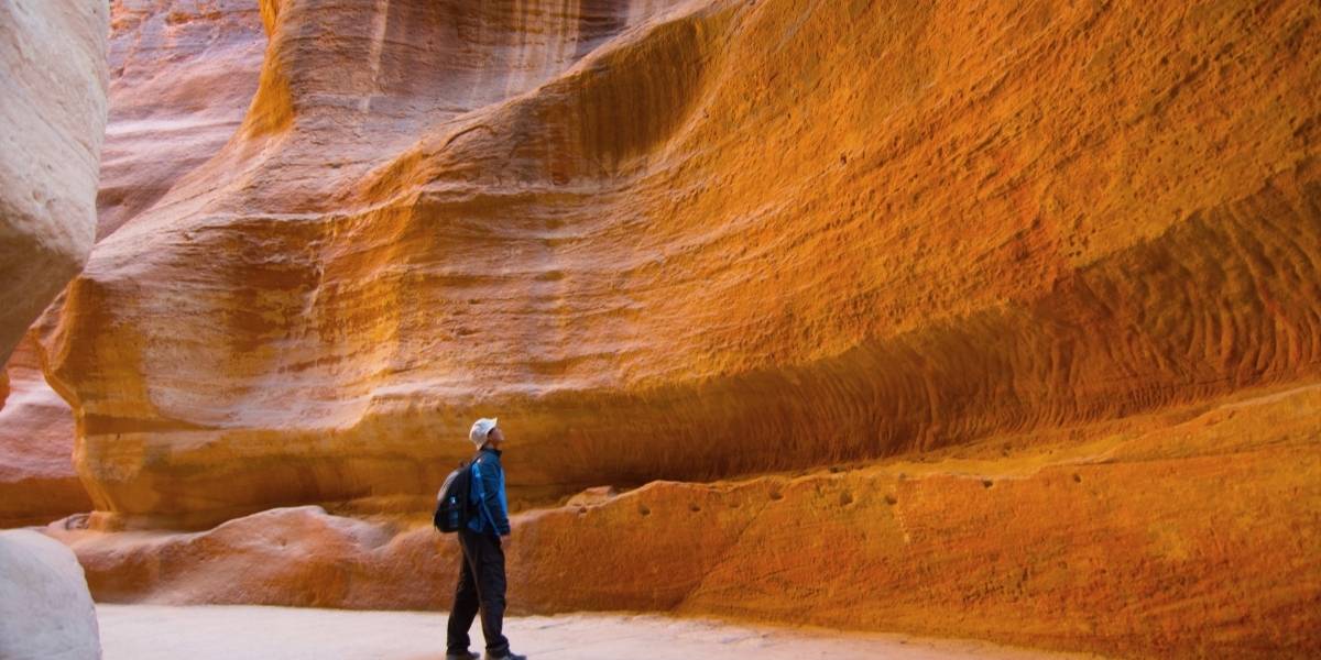 Hiker wearing a blue jacket and backpack stands in a narrow orange sandstone canyon of layered rock formations.