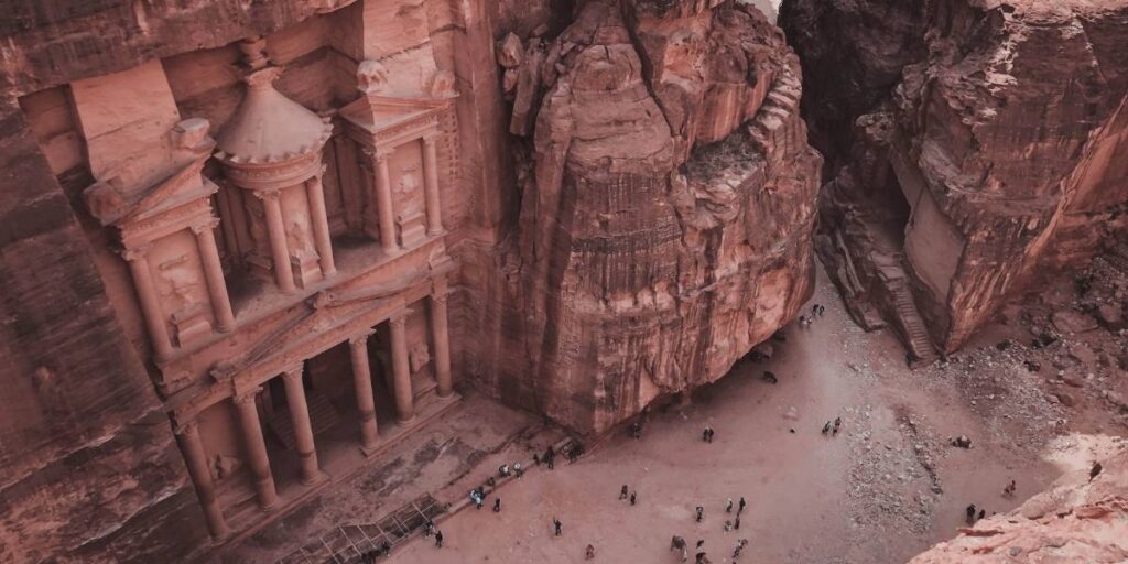 Aerial view of the Khazneh temple carved into a red sandstone cliff at Petra, with visitors at the sandy yard below.