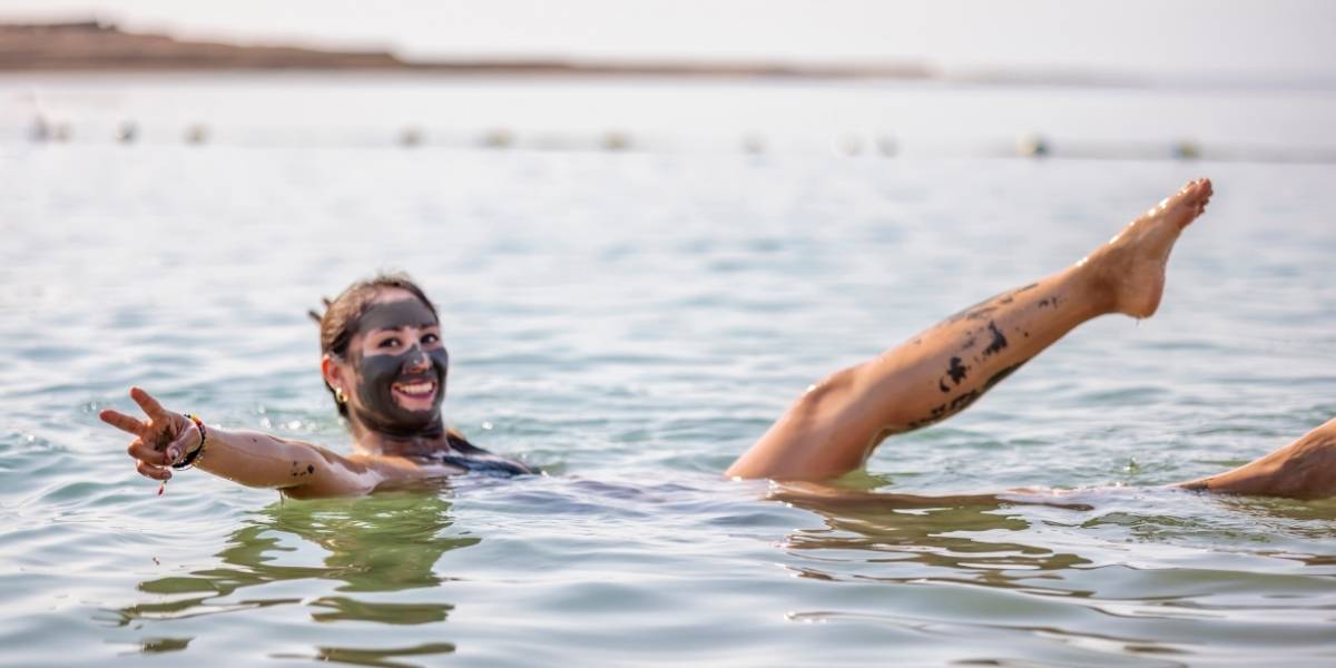 Woman in a calm sea smiling and waving with a peace sign, wearing a dark facial mud mask and visible leg tattoos.