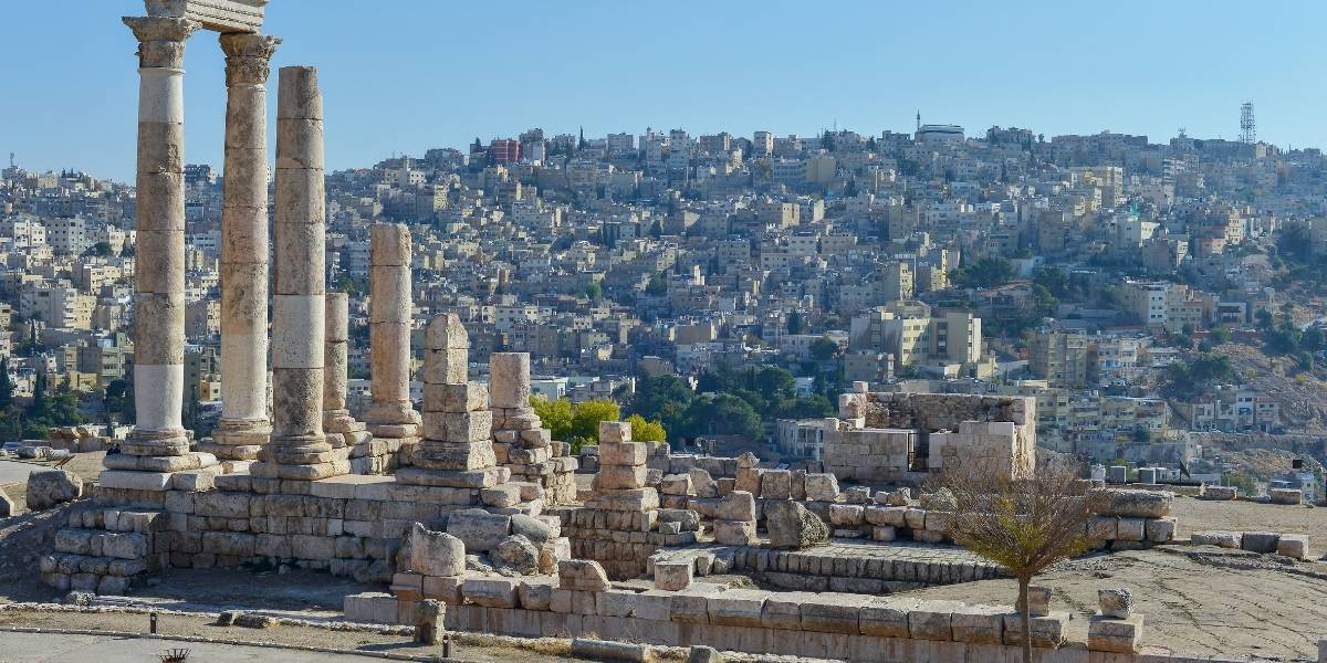 Ancient stone columns and ruins in the foreground with a densely built hillside city in the background, under a clear blue sky.