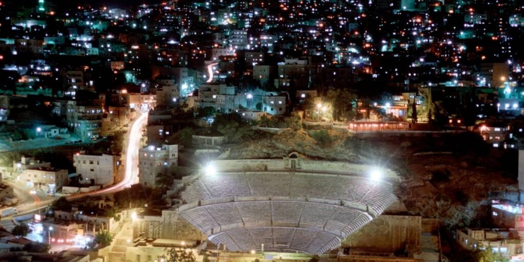 Nighttime city view with an ancient outdoor amphitheater in the foreground and illuminated streets behind.