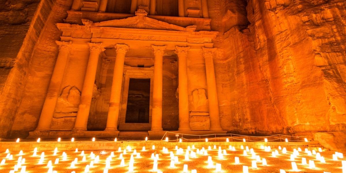 Petra’s rock‑cut temple facade illuminated by orange light with many candles on the ground in front of stairs and columns.