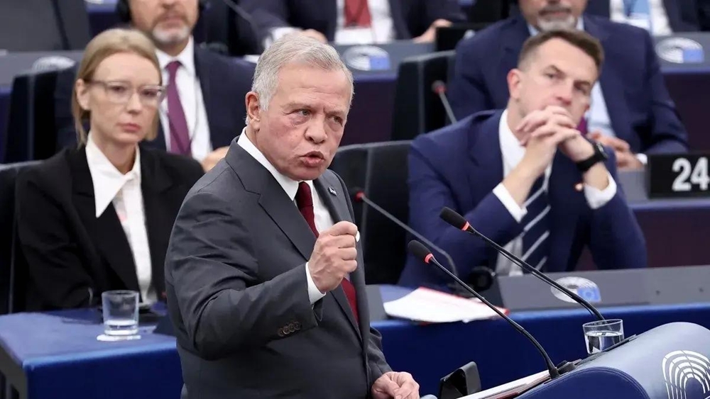 Man in a gray suit and red tie speaks passionately at a podium with microphones in a formal conference setting.