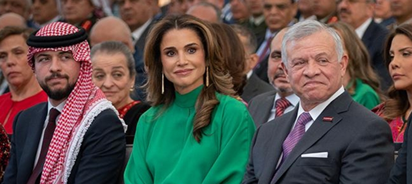 Group of formally dressed attendees seated at a public event; woman in a green blouse at center flanked by two men in suits, outdoors
