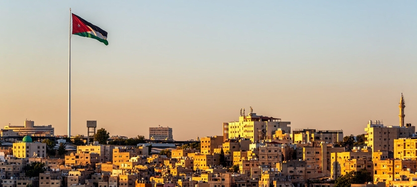 Sunlit Amman cityscape at sunset with a tall flagpole flying the Jordan flag over dense apartment buildings.