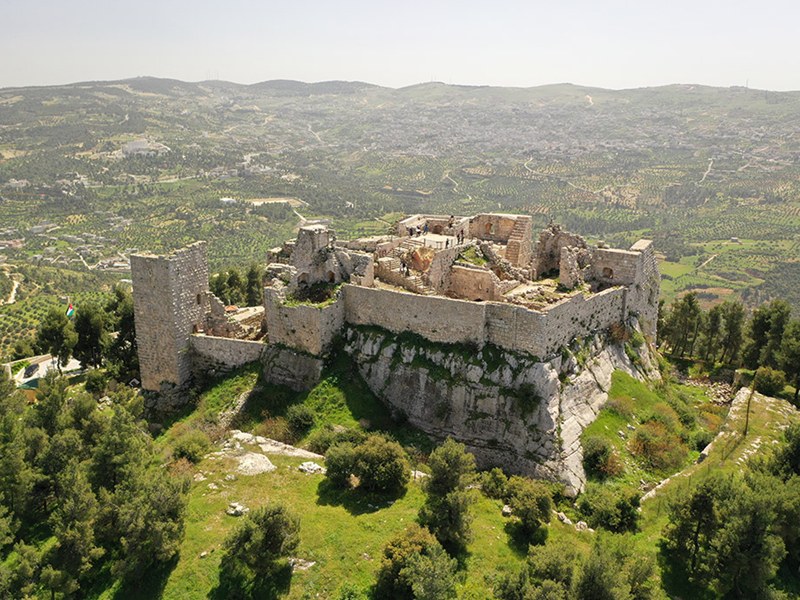 Ruined medieval castle atop a rocky hill overlooking a green valley and distant hills.