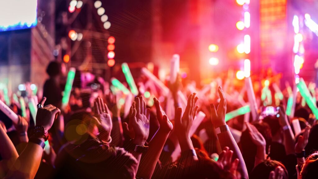 Crowd at an energetic concert with raised hands and colorful stage lights in pinks, purples, and greens.