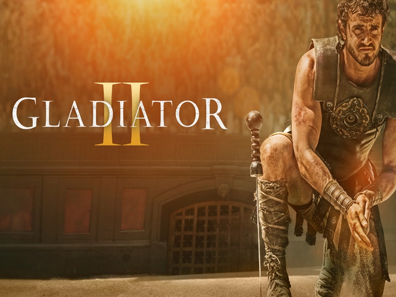 Promotional image for Gladiator II featuring a battle-worn gladiator in armor against a dusty arena backdrop with warm orange light.