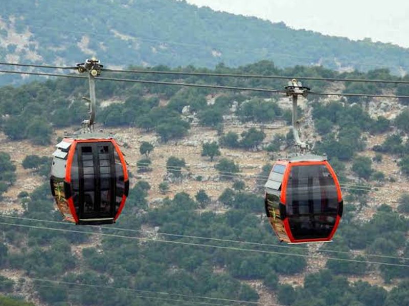 Two red gondola cabins travel along a mountain cable car line over a forested hillside.