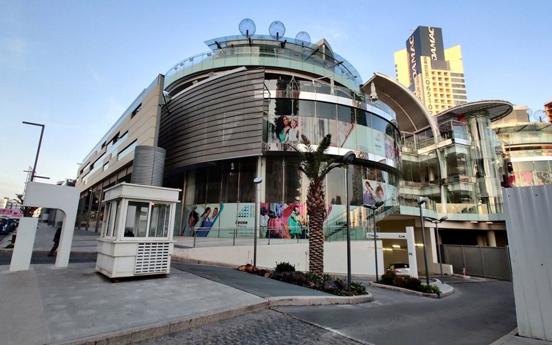 Curved glass shopping center with palm tree, storefront banners, and a pedestrian area in front