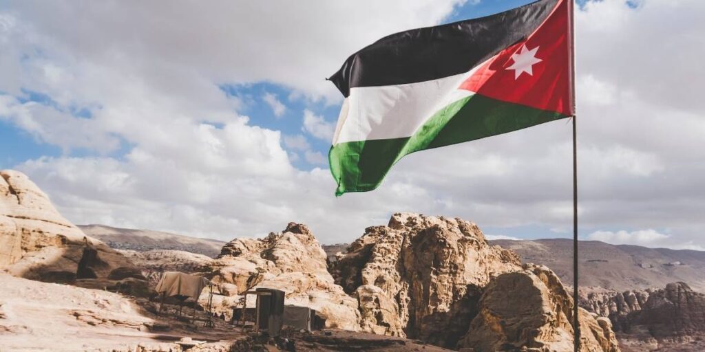 Jordanian flag fluttering over a rocky desert campsite with tents beneath a blue sky.