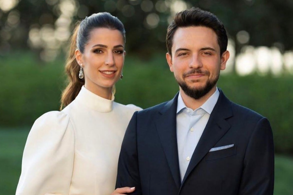Couple dressed formally posing outdoors; woman in white high-neck dress with pearl earrings, man in navy suit and light blue shirt.