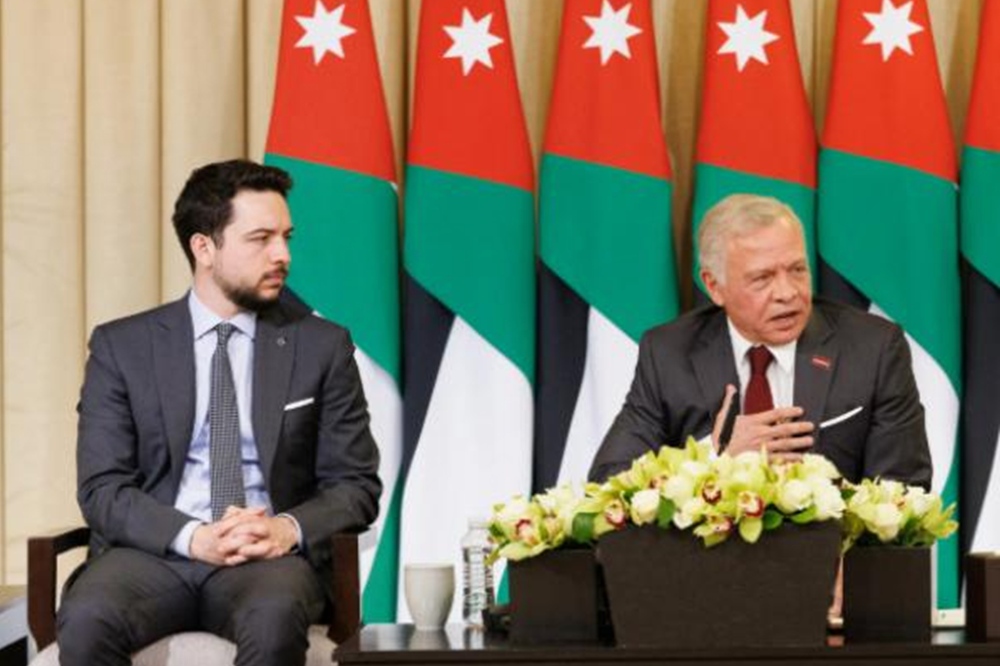 Two men in suits at a formal press conference; one speaks from a flower‑decorated podium while the other listens, with Jordanian flags in the background.