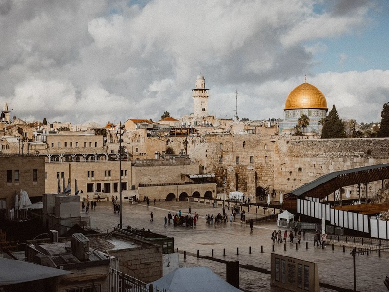 Temple Mount plaza in Jerusalem with the Dome of the Rock's gold dome and ancient limestone walls, people walking on a wet stone square under a cloudy sky