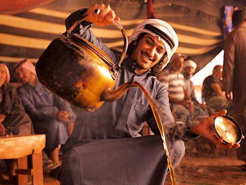 Man in traditional dress pours tea from a brass kettle into a cup for guests in a crowded tented area with smiling onlookers behind him.