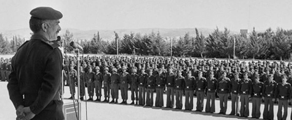 Senior officer in a beret speaks into a microphone to a long line of uniformed soldiers standing at attention outdoors.