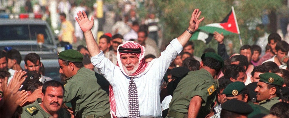 Man with a red-and-white keffiyeh raises both arms in a crowded political rally, surrounded by uniformed soldiers; a flag waves in the background.