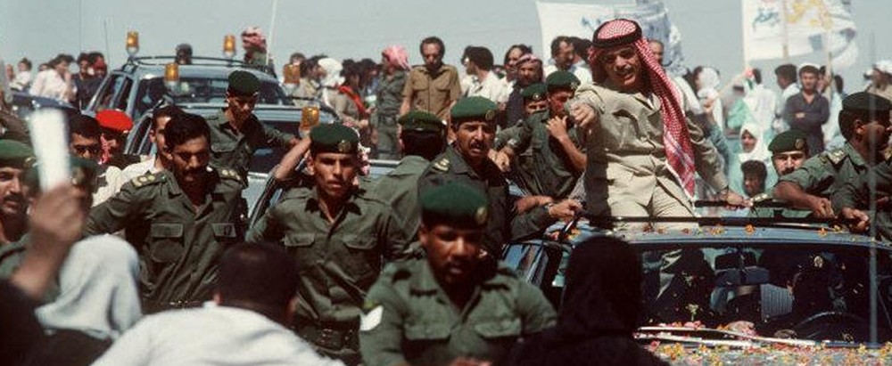 Crowd surrounding a decorated convertible as uniformed security personnel escort a man wearing a red-and-white keffiyeh, waving from the car.