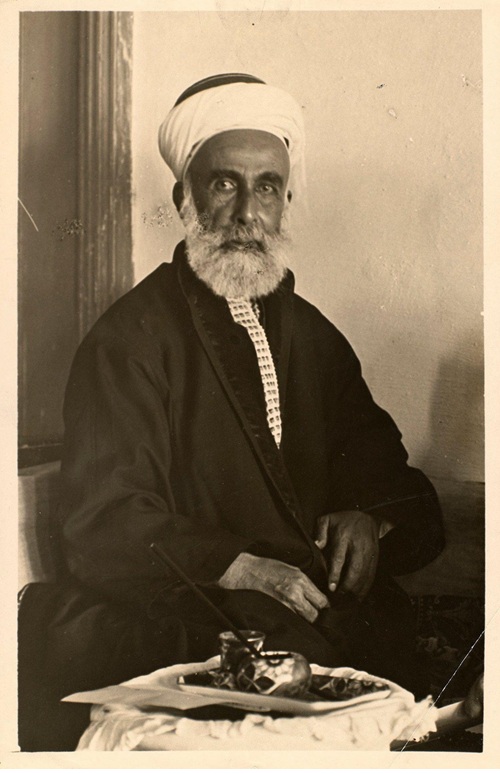 Portrait of an elderly man with a white beard, wearing a turban and dark robe, seated with a tea service on a table nearby.