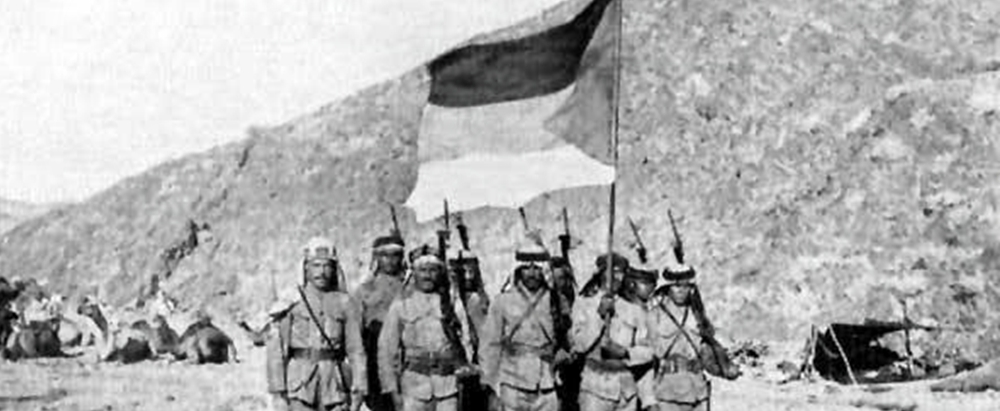 Line of soldiers marching with rifles and bayonets, carrying a tall flag across a barren desert scene.