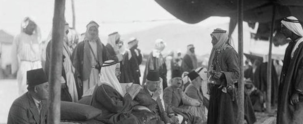 Group of men in traditional robes and headdresses gathered under tents in a desert camp, listening to a speaker