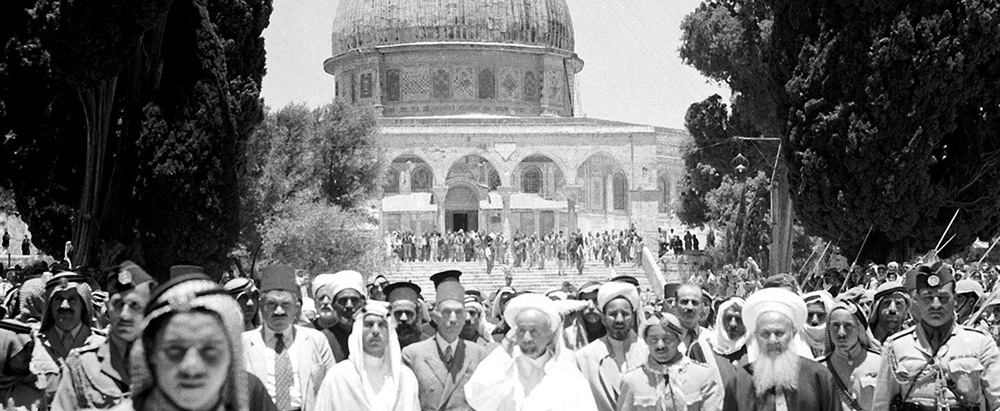 Large crowd in traditional dress marching toward a domed shrine in Jerusalem, with trees on the sides.