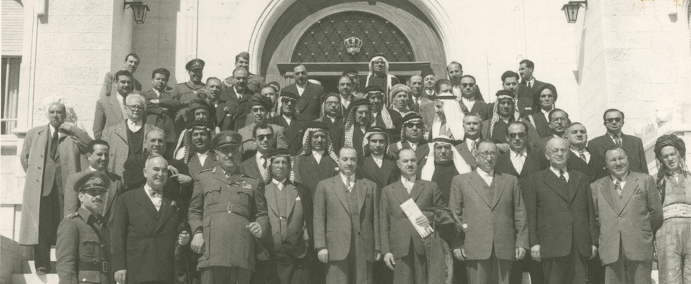 Large group of men in suits, traditional dress, and a military officer posing on the steps of an official building.
