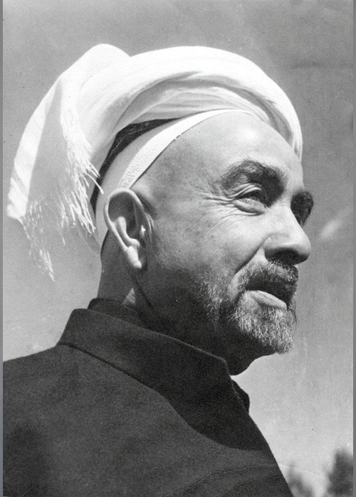 Side-profile of a man wearing a white turban and dark coat, looking into the distance.