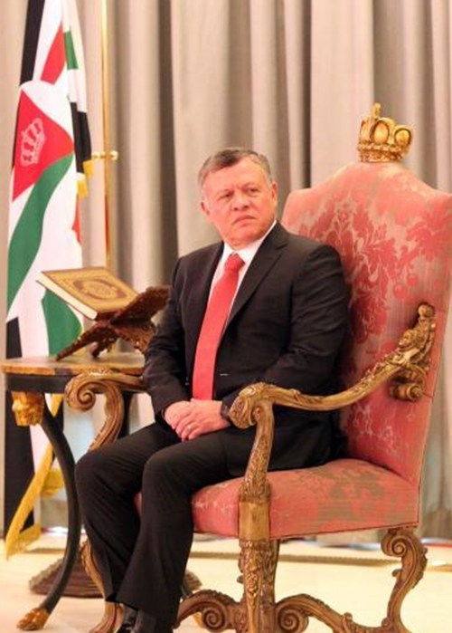 A formally dressed man in a black suit and red tie sits on an ornate pink and gold throne, with a Palestinian flag in the background.