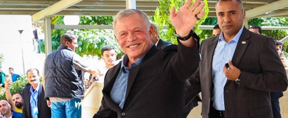 Smiling older man in a dark suit waves to the camera, surrounded by supporters outdoors under a shaded walkway.