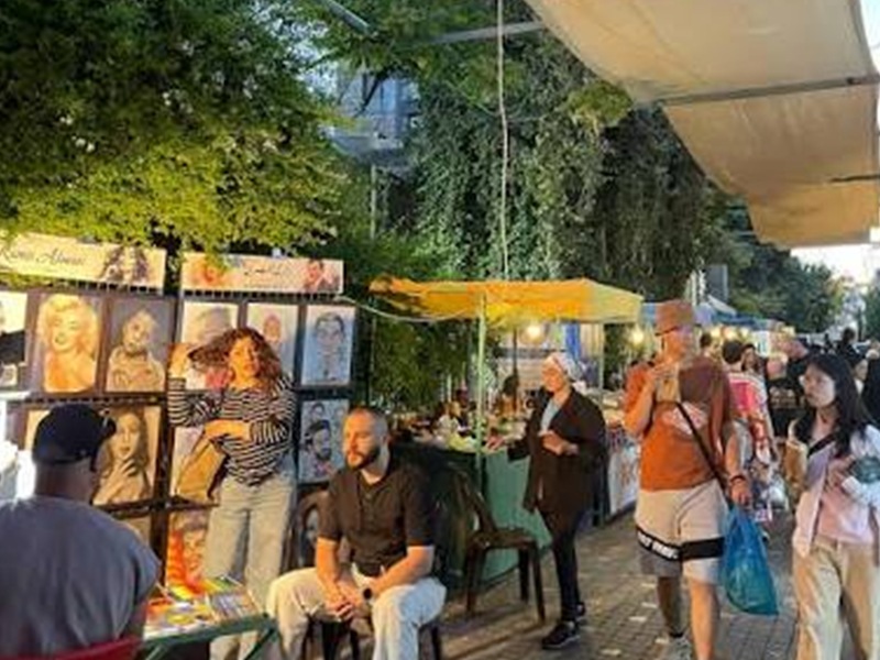 Outdoor street market with artist stalls displaying portraits; people browse and chat under colorful tents as evening light falls.