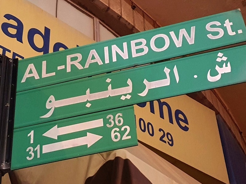 Green street sign reading 'AL-RAINBOW St.' with Arabic text, featuring white arrows and route numbers 1, 31 to the left and 36, 62 to the right; yellow signs in the background.