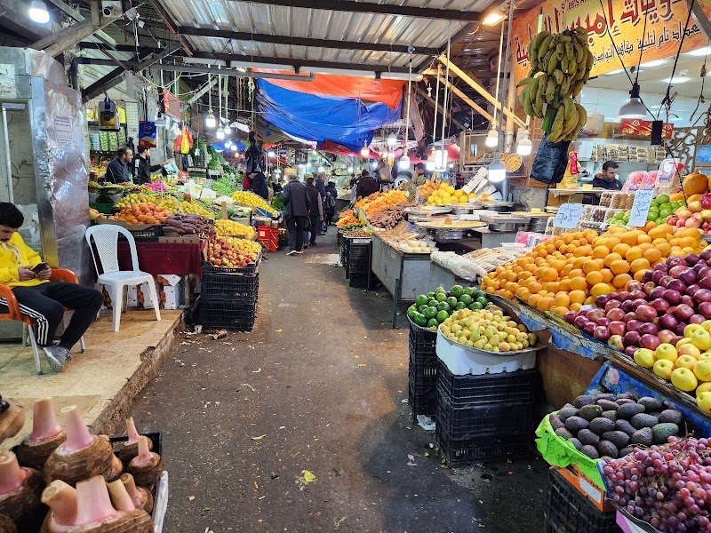 Busy indoor market scene with fruit stalls on both sides, vendors and shoppers among baskets of oranges, apples, bananas, and root vegetables under colorful canopies.
