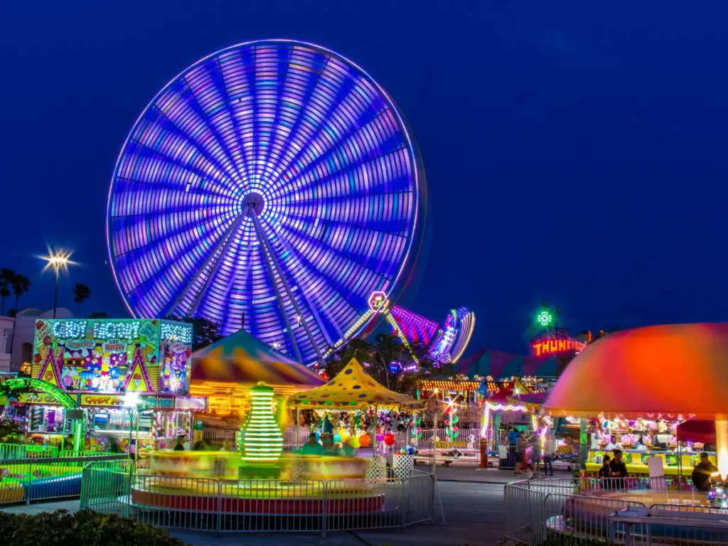 Night carnival at dusk with a large, illuminated blue-purple Ferris wheel and colorful stalls and rides glowing in neon lights at the fairgrounds.