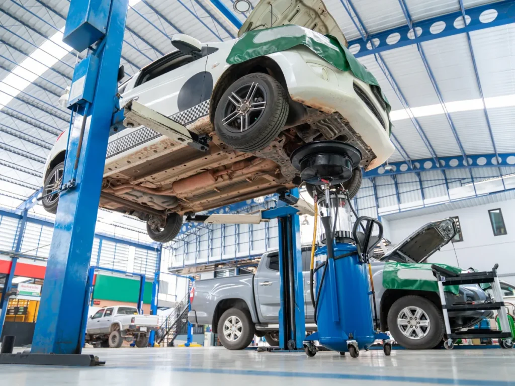 Car raised on a two-post hydraulic lift in a bright auto repair shop, awaiting service with the undercarriage visible and tools nearby.